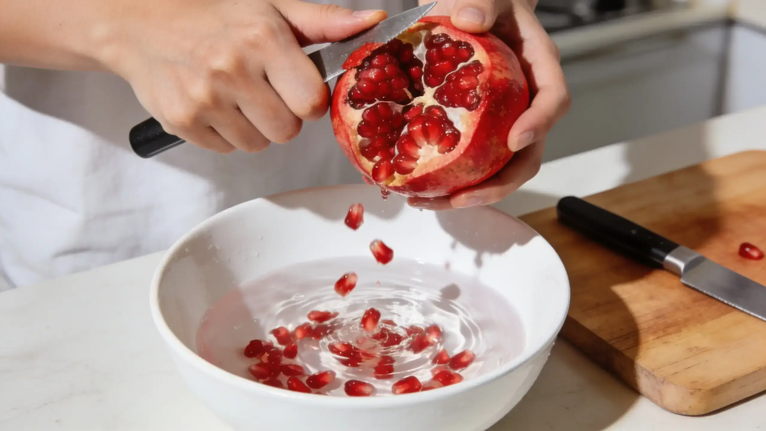Grenade : 8 Bienfaits Santé du Fruit et Comment la Manger 2 Mains épépinant une grenade ouverte au-dessus d'un bol d'eau blanc avec les arilles rouges qui tombent dans l'eau, couteau et planche à découper en bois visibles à côté sur plan de travail clair