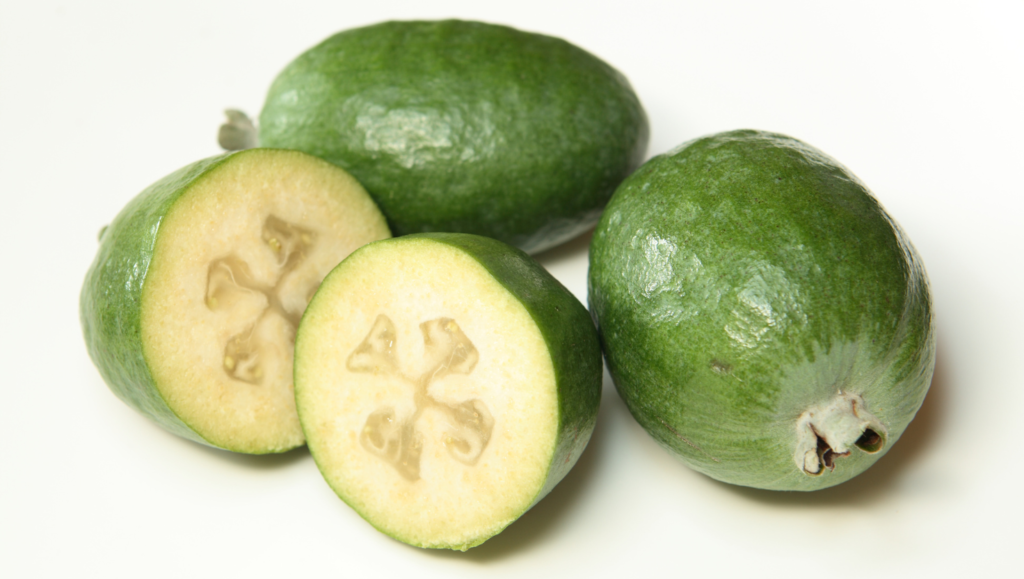 Feijoas verts posés sur une surface blanche, avec deux fruits coupés en deux montrant leur chair beige translucide et leurs graines en forme d’étoile.