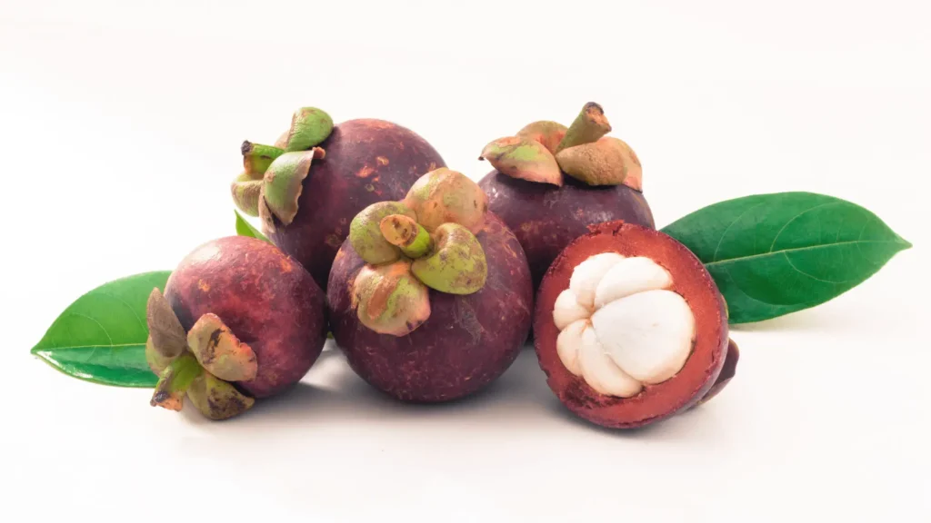 Plusieurs mangoustans violets avec leur calice vert, dont un coupé en deux montrant sa chair blanche segmentée, posés sur un fond blanc avec des fe
