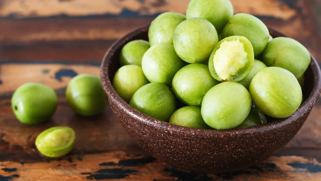 Bol rempli de petits fruits verts umbus, avec un fruit croqué révélant sa chair jaune juteuse, posé sur une table en bois.