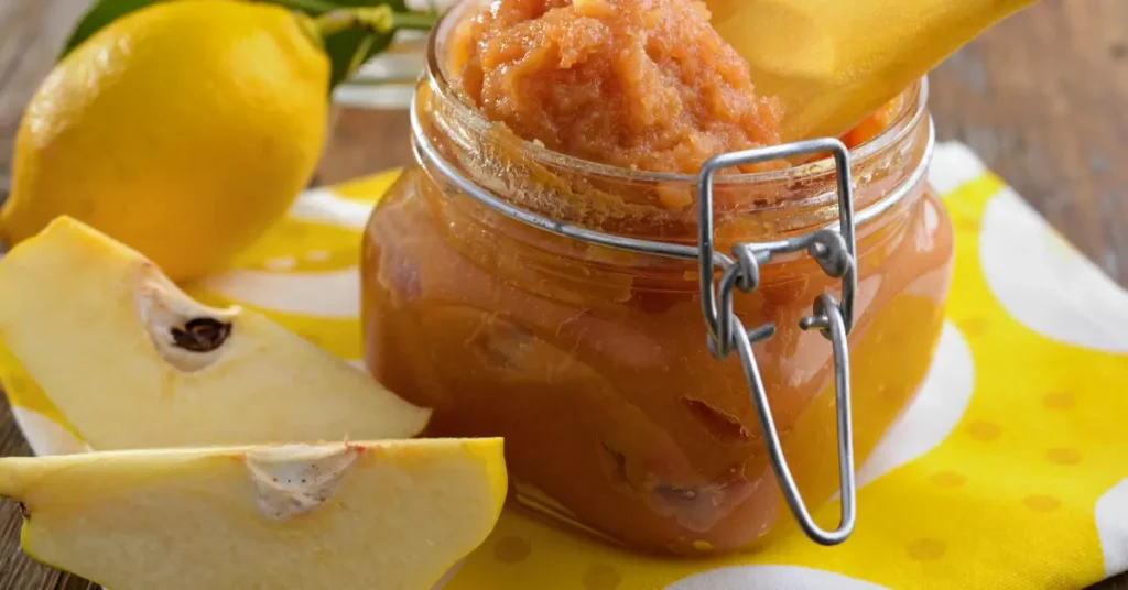 Pot en verre de confiture de coings avec cuillère en bois, entouré de tranches de coing frais et d’un citron sur un torchon jaune à pois blancs.