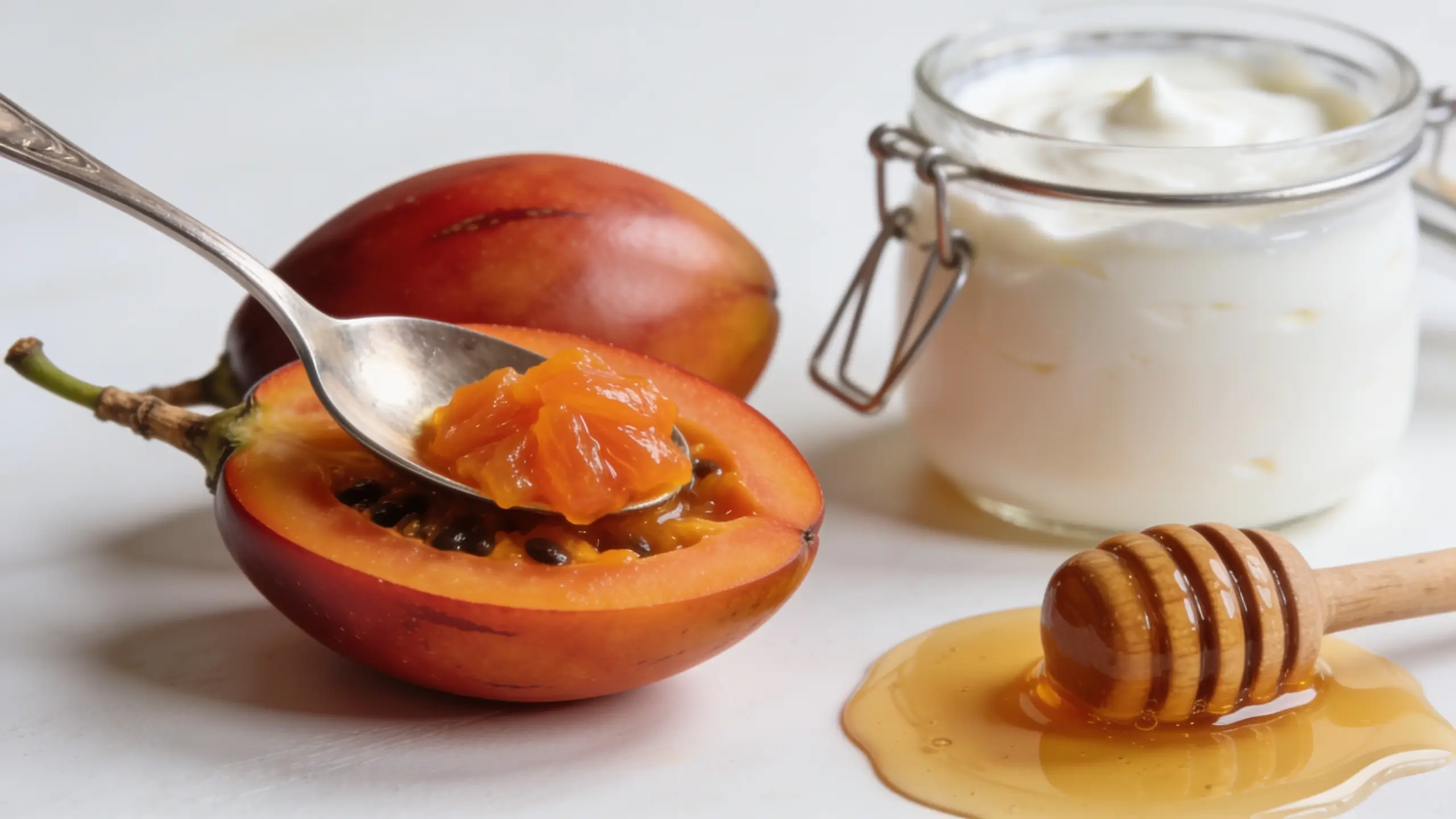 Cuillère prélevant la chair orange d'un tamarillo coupé en deux posé sur fond blanc, avec un pot de yaourt grec et un filet de miel à côté, façon de manger le tamarillo à la cuillère