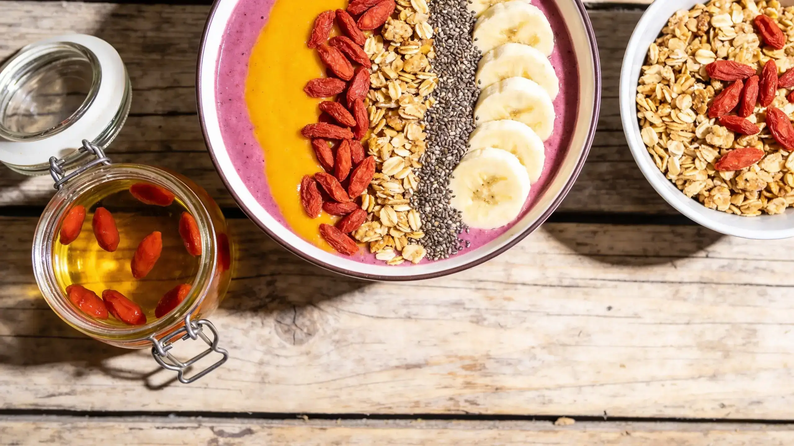 Baie de Goji Bienfaits : 7 Secrets Santé Révélés 5 Smoothie bowl bicolore jaune et rose garni de baies de goji, granola, graines de chia et tranches de banane sur table en bois, avec bocal de baies de goji au miel à côté